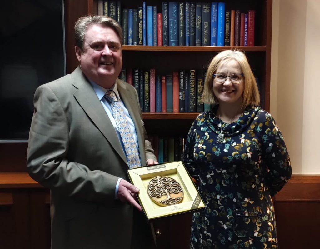 Chairperson of Twilight Community Group, Martin Brennan, holds placard of tree. Martin Brennan and Polish Ambassador to Ireland, HE Anna Sochańska, smile to camera. Both are posing in front of bookshelf.